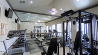 This fitness center offers a thoughtfully designed workout environment suited for a wide range of exercise routines. The space is lined with large floor-to-ceiling mirrors along one wall, creating a sense of expansiveness and allowing users to monitor their form during workouts. Recessed lighting and a ceiling fan provide ample illumination and airflow throughout the room, contributing to a comfortable training atmosphere.

The facility is well-stocked with a variety of equipment to support both cardiovascular and strength training goals. Cardio machines, including treadmills and other aerobic equipment, are positioned near the windows, allowing natural light to filter in through the blinds. A multi-function cable and weight machine anchors the strength training area, complemented by flat weight benches and free weights neatly arranged on floor mats. The neutral-toned walls and light flooring lend a clean, functional aesthetic to the space.

Overall, this fitness center strikes a balance between practicality and comfort, providing users with the tools and environment needed for an effective workout. The combination of natural light, mirrored walls, and a diverse equipment selection makes this a versatile and inviting space for fitness enthusiasts of all levels.