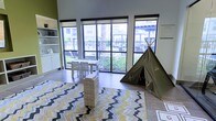 This single-floor activity space is designed to inspire creativity and play in a bright, welcoming environment. Large floor-to-ceiling windows line the walls, flooding the interior with abundant natural light and offering views of lush outdoor greenery, palm trees, and surrounding landscaping. The open layout is anchored by light hardwood-style flooring, which complements the space's airy, modern aesthetic.

A standout feature of the room is a bold chevron-patterned area rug in shades of yellow, blue, gray, and white, which defines the central play zone and adds a lively burst of color. A canvas teepee tent in an earthy olive green serves as a charming focal point, inviting imaginative play. A stacked wooden block tower game adds an element of interactive fun to the space.

A small white table with matching chairs is positioned near the windows, providing a functional area for seated activities. Built-in white shelving and cabinetry along the walls offer organized storage and display space. Neutral-toned walls with a subtle accent of green enhance the room's fresh, playful character.

Overall, the space strikes a balance between functional and fun, making it a well-appointed communal area suited for leisure, play, and social interaction in a naturally lit, cheerful setting.