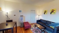 This single-floor space offers a warm and inviting atmosphere defined by classic hardwood flooring and notably high ceilings that lend an open, airy quality to the interior. The living area is thoughtfully arranged, featuring a dark leather sofa paired with a tufted ottoman and a wooden coffee table, all anchored by a decorative area rug with rich, patterned detailing. A floor lamp provides ambient lighting, complementing the clean white walls that serve as a neutral backdrop throughout the space. A paneled closet door adds a traditional architectural touch, while practical storage solutions blend seamlessly into the room. Colorful artwork displayed on the walls adds personality and character to the otherwise understated palette. The layout makes efficient use of the available space, balancing functional storage with comfortable living areas. The combination of classic architectural elements — including tall baseboards and the warm tones of the hardwood floors — creates a cozy yet timeless ambiance suited to a variety of lifestyles.
