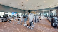 Located at 1605 Barclay Point Blvd in Wilmington, NC, this single-floor fitness center is a comprehensively outfitted facility designed to support a wide range of workout routines. The space is defined by deep teal-blue walls, wood-style flooring, and recessed lighting complemented by pendant ceiling fans, creating a modern and energizing atmosphere throughout.

The cardio area features a row of Life Fitness treadmills positioned in front of large, white-framed windows that invite generous natural light and offer pleasant outdoor views of the surrounding greenery. Nearby, a dedicated zone houses elliptical cross-trainers and wall-mounted flat-screen televisions, making endurance workouts both comfortable and engaging.

The strength training floor is generously appointed with a comprehensive selection of Life Fitness selectorized machines, free weights, benches, and additional resistance equipment. Full-length mirrors along the walls expand the sense of space and allow users to monitor their form during workouts.

A glass-partitioned cycling studio provides a dedicated space for spin workouts, featuring LifeCycle bikes and a frosted-glass accent wall that lends the room a polished, boutique feel. The studio connects visually to the main floor through floor-to-ceiling glass panels, maintaining an open and cohesive feel across the entire facility.