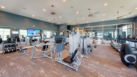 Located at 1605 Barclay Point Blvd in Wilmington, NC, this single-floor fitness center is a comprehensively outfitted facility designed to support a wide range of workout routines. The space is defined by deep teal-blue walls, wood-style flooring, and recessed lighting complemented by pendant ceiling fans, creating a modern and energizing atmosphere throughout.

The cardio area features a row of Life Fitness treadmills positioned in front of large, white-framed windows that invite generous natural light and offer pleasant outdoor views of the surrounding greenery. Nearby, a dedicated zone houses elliptical cross-trainers and wall-mounted flat-screen televisions, making endurance workouts both comfortable and engaging.

The strength training floor is generously appointed with a comprehensive selection of Life Fitness selectorized machines, free weights, benches, and additional resistance equipment. Full-length mirrors along the walls expand the sense of space and allow users to monitor their form during workouts.

A glass-partitioned cycling studio provides a dedicated space for spin workouts, featuring LifeCycle bikes and a frosted-glass accent wall that lends the room a polished, boutique feel. The studio connects visually to the main floor through floor-to-ceiling glass panels, maintaining an open and cohesive feel across the entire facility.