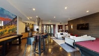 Located at 6715 E Union Ave in Denver, CO, this single-floor amenity lounge is thoughtfully designed to serve as a premier social and entertainment hub. The open-concept layout is anchored by polished concrete floors that reflect recessed lighting, lending the space a sleek, contemporary character throughout.

The kitchenette area is outfitted with warm bamboo cabinetry, dark stone countertops, a stainless steel dishwasher, a commercial beverage station, and a reflective tile backsplash that adds visual depth. A round high-top table with leather bar stools nearby provides a casual spot for conversation or dining.

The main lounge area features a generously sized seating arrangement with a cream-toned sectional sofa, red accent ottomans, and a flat-screen TV mounted on a striking wood-paneled feature wall illuminated by modern sconce lighting. Adjacent to the lounge sits a regulation-size billiards table with a bold red felt surface, positioned beneath pendant lighting and beside a large window that draws in natural light and offers views of an outdoor patio area.

A semi-private nook with a round walnut table, leather chairs, a floor-length mirror, and original artwork provides an intimate setting for smaller gatherings. Curated sculptures and paintings are displayed throughout, elevating the overall ambiance of this well-appointed communal space.