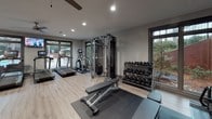 This fitness center is designed to support a comprehensive workout experience within a single, open-plan floor. The space features light wood-style flooring throughout, neutral gray walls, and recessed lighting complemented by a ceiling fan, creating a clean and energizing atmosphere. Large windows along multiple walls invite natural light and offer views of surrounding greenery and an adjacent pool area, adding a refreshing outdoor connection to the interior environment.

The cardio zone is well-appointed with multiple treadmills, an elliptical trainer, a stair climber, and stationary cycling bikes — several equipped with digital displays for interactive workouts. A wall-mounted television provides entertainment during cardio sessions. The strength training area includes a multi-station cable machine, a full rack of dumbbells, a tiered kettlebell rack with a colorful assortment of weights, an adjustable workout bench, and a stability ball rack. Three large black-framed mirrors line one wall, enhancing the sense of space and allowing users to monitor their form during exercise. Rubber floor mats are placed beneath equipment stations for safety and performance. Abstract wall art adds a touch of character to the space. The overall layout is thoughtfully organized, offering clear zones for cardio, strength, and functional training without feeling overcrowded.