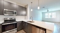 This single-floor residence showcases a clean, contemporary design throughout. The kitchen is a standout feature, equipped with dark wood-grain cabinetry, white quartz countertops, and a full suite of stainless steel appliances including a refrigerator, electric range, over-the-range microwave, and dishwasher. A peninsula with an undermount sink provides additional workspace and serves as a natural transition into the open living area. Stylish pendant lighting with exposed bulbs hangs above, adding a modern industrial touch. The living area is bright and open, with light wood-look flooring that extends throughout the main spaces, crisp white walls, and a window that invites natural light. A built-in desk nook adds a practical workspace element near the entryway. The bedroom features plush carpeting, a ceiling fan, and a window with blinds offering outdoor views and natural light. The bathroom is generously sized with wood-look flooring, a dark-toned vanity with white countertop, a framed mirror, and direct access to a walk-in closet with built-in shelving and hanging rods. Additional open shelving near the bathroom entry provides convenient storage. The overall ambiance is fresh, modern, and move-in ready.