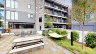 This inviting outdoor courtyard is thoughtfully designed to provide residents with a comfortable and functional retreat. Set within the interior of a contemporary multi-story building, the space benefits from a sheltered feel while remaining open to natural light and the surrounding greenery. The courtyard's modern aesthetic is reflected in the building's exterior, which combines dark horizontal siding, stone accents, and large windows that overlook the shared amenity area.

At the heart of the courtyard, a bright yellow hammock suspended between posts serves as a standout focal point, offering a casual spot for relaxation. Nearby, a cluster of Adirondack chairs arranged on a stone-paved patio provides an additional gathering area ideal for conversation or quiet leisure. Picnic-style tables with bench seating offer a practical space for outdoor dining or socializing.

Lush, manicured shrubs and mature trees are woven throughout the courtyard, softening the hardscape and contributing to a green, park-like atmosphere. Stepping stone pathways connect the various seating areas, guiding visitors through the space with ease. The combination of thoughtful landscaping, varied seating options, and contemporary architectural surroundings creates a welcoming communal environment suited for everyday outdoor enjoyment.