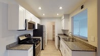 This single-floor kitchen is designed for both functionality and style, offering a thoughtfully organized galley-style layout. White shaker-style cabinetry lines both sides of the space, providing generous storage while contributing to a clean, bright aesthetic. The dark granite countertops add contrast and a touch of elegance, complementing the stainless steel appliance suite, which includes a gas range with overhead microwave, a refrigerator, and a dishwasher. Recessed lighting is installed throughout the ceiling, ensuring consistent, well-distributed illumination across the entire workspace. The double-basin stainless steel sink is positioned along one counter run, paired with a modern gooseneck faucet. Light wood-toned flooring runs the length of the kitchen, adding warmth to the overall palette. A standout feature is the walk-in pantry accessible directly from the kitchen, fitted with open shelving that offers practical dry storage. At the far end, a glimpse of an adjacent room hints at a connected, open living arrangement. Overall, the kitchen balances practical design with contemporary finishes.