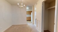 Located at 300 Hidenwood Place in Newport News, Virginia, this single-floor residence offers a well-organized and functional layout designed for comfortable everyday living. The dining area makes an immediate impression with its elegant chandelier, featuring a classic candelabra-style design with brass accents that adds a refined touch to the space. Neutral-toned carpeting runs throughout, creating a warm and cohesive feel underfoot. The dining area flows seamlessly into the kitchen, which is outfitted with warm wood-toned cabinetry offering ample storage, light-colored countertops, and a full-size refrigerator. The kitchen flooring transitions to a smooth, easy-to-clean surface, providing a practical contrast to the carpeted living areas. Louvered interior doors contribute a classic architectural detail while allowing for ventilation throughout the space. The light, neutral color palette across walls and floors creates a bright and airy ambiance that makes the interior feel open and welcoming. Overall, this single-story residence at Hidenwood Place combines straightforward design with functional features, making it a practical and comfortable home in the Newport News area.