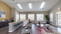 This single-floor community space is thoughtfully designed to serve as both a welcoming social hub and a functional leasing or management office. The architecture is defined by soaring vaulted ceilings that lend an airy, open feel throughout, complemented by neutral gray walls and rich dark hardwood-style flooring that flows seamlessly from room to room.

The main lounge area is generously sized, featuring a stylish seating arrangement with a gray sectional sofa, patterned accent chairs, and a vibrant pink geometric area rug. Multiple large windows dressed with white horizontal blinds fill the space with abundant natural light and offer views of the surrounding greenery. Elegant glass-top coffee tables and coordinated décor complete the inviting atmosphere.

Adjacent to the lounge, a reception area is anchored by a substantial dark wood desk, framed artwork, and direct sightlines to the entrance — ideal for staff-resident interaction. French doors with grid-pane glass panels connect different zones of the space, enhancing both openness and architectural character.

A dedicated dining nook features a round table with modern chairs set against a warm golden accent wall, alongside a beverage station with a mini-refrigerator and coffee maker. The kitchenette area is outfitted with granite countertops, dark cabinetry, a stainless steel sink, and ample counter space, providing practical amenity support for the overall facility.
