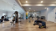 This spacious fitness center is thoughtfully designed to support a full range of workout routines across a single, open floor plan. The space features warm hardwood flooring throughout, neutral-toned walls, and recessed lighting complemented by ceiling fans, creating a bright and comfortable environment for exercise.

The cardio zone is well-appointed with multiple treadmills, elliptical trainers, stationary bikes, and rowing machines, all positioned near large windows that invite natural light and outdoor views. A wall-mounted television keeps users entertained during cardio sessions.

The strength training area offers a diverse selection of cable and weight machines, adjustable benches, and a dedicated free weights section with a full rack of dumbbells. Large floor-to-ceiling mirrors line the walls of this zone, enhancing the sense of openness while allowing users to monitor their form.

A functional training area houses medicine balls, stability balls, and suspension training equipment, offering versatility for dynamic workouts. A wall-mounted air conditioning unit ensures climate control throughout the space.

The overall ambiance is clean, modern, and professional, with generous spacing between equipment that allows for comfortable movement. The combination of natural light, reflective mirrors, and quality flooring gives this fitness center a polished, gym-quality feel suited for a broad range of fitness needs.