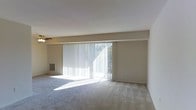 This single-story space features a spacious, open living area designed with simplicity and functionality in mind. The room is finished with neutral-toned carpeting that extends throughout, complementing the clean white walls and ceiling. A prominent architectural beam runs horizontally across the upper portion of the far wall, adding subtle structural definition to the space.

The most notable feature of the living area is the large sliding glass door spanning a significant portion of the exterior wall. Dressed with vertical blinds, this door allows abundant natural light to flood the interior, casting bright patterns across the floor and creating an airy, open atmosphere. The door also provides direct access to the outdoors, enhancing the sense of connection between the interior and exterior.

A ceiling fan is positioned overhead, offering practical climate control and air circulation throughout the room. A floor-level air vent is also visible along the baseboard, indicating a built-in heating and cooling system. The overall design is clean and understated, offering a versatile, move-in-ready environment well-suited for a variety of furniture arrangements and personal styles.