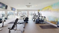 The fitness center at 2901 4th Ave S in Birmingham, Alabama is a well-appointed, single-floor facility designed to support a comprehensive workout experience. The space is bathed in natural light, courtesy of large floor-to-ceiling windows along multiple walls that frame sweeping views of the Birmingham cityscape, creating an energizing atmosphere for exercise. The interior features durable, cushioned flooring throughout, providing comfort and safety for a variety of activities. A striking abstract mural in soft blues, yellows, and greens serves as a bold focal point, adding a vibrant artistic element to the otherwise clean, white-walled space. The cardio section is well-equipped with multiple Matrix treadmills, elliptical machines, stationary bikes, and air resistance bikes, all positioned to take advantage of the scenic outdoor views. A dedicated strength training area includes adjustable weight benches, a rack of dumbbells, and a cable machine, reflected in a large wall-mounted mirror that enhances the sense of openness. A wall-mounted flat-screen television provides entertainment during workouts. The facility also features a cassette-style HVAC unit in the ceiling for climate control and direct access to an adjacent outdoor terrace, seamlessly extending the fitness experience outdoors.