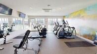 The fitness center at 2901 4th Ave S in Birmingham, Alabama is a well-appointed, single-floor facility designed to support a comprehensive workout experience. The space is bathed in natural light, courtesy of large floor-to-ceiling windows along multiple walls that frame sweeping views of the Birmingham cityscape, creating an energizing atmosphere for exercise. The interior features durable, cushioned flooring throughout, providing comfort and safety for a variety of activities. A striking abstract mural in soft blues, yellows, and greens serves as a bold focal point, adding a vibrant artistic element to the otherwise clean, white-walled space. The cardio section is well-equipped with multiple Matrix treadmills, elliptical machines, stationary bikes, and air resistance bikes, all positioned to take advantage of the scenic outdoor views. A dedicated strength training area includes adjustable weight benches, a rack of dumbbells, and a cable machine, reflected in a large wall-mounted mirror that enhances the sense of openness. A wall-mounted flat-screen television provides entertainment during workouts. The facility also features a cassette-style HVAC unit in the ceiling for climate control and direct access to an adjacent outdoor terrace, seamlessly extending the fitness experience outdoors.
