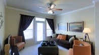 This single-floor space offers a thoughtfully designed interior that balances comfort and style. The living room serves as the centerpiece, showcasing elegant crown molding that frames the ceiling alongside a classic five-blade ceiling fan with integrated lighting. Large windows dressed with dramatic dark curtains allow generous natural light to fill the room while offering the option for full privacy. A set of glass doors leads to an outdoor balcony, seamlessly connecting the interior to the outside. The space is finished with plush carpeting underfoot, contributing to a warm and inviting atmosphere. Furnishings are tastefully arranged, with a deep brown sofa accented by earth-toned throw pillows, a substantial upholstered ottoman doubling as a coffee table, and coordinating accent chairs in a warm caramel hue. Framed artwork adorns the walls, adding a polished, curated feel to the overall aesthetic. The neutral wall tones provide a versatile backdrop that complements the rich, layered color palette of the furnishings. Together, these elements create a cohesive living environment that feels both functional and refined, ideal for everyday living and entertaining alike.