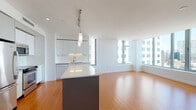 Located on Essex Street in Boston's dynamic downtown district, this modern single-floor residence showcases a clean, contemporary design throughout. Warm bamboo hardwood flooring flows seamlessly across the open-plan living and dining areas, complementing the bright white walls and crisp architectural lines.

The kitchen is a standout feature, equipped with stainless steel appliances including a range, microwave, and refrigerator. Flat-front white cabinetry extends to the ceiling, paired with light quartz countertops, a subway tile backsplash, and a deep stainless steel sink. A central island with a contrasting dark base provides additional prep space and serves as a natural gathering point.

The living area is generously proportioned, with large floor-to-ceiling windows framing sweeping views of Boston's downtown skyline, filling the space with abundant natural light. The bedrooms continue the bamboo flooring and feature large windows with impressive urban vistas.

The bathrooms are finished in a modern style, featuring large-format neutral floor tiles, floating dark-wood vanities with white countertops, backlit mirrors, and clean white walls. The overall ambiance of the residence is bright, minimalist, and polished, well-suited to urban living in the heart of Boston.