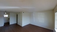 This single-floor space offers a clean, functional layout designed for comfortable everyday living. The main living area is highlighted by rich, dark hardwood-style flooring that contrasts elegantly with the bright, neutral white walls, creating a warm yet modern aesthetic. A textured ceiling adds subtle visual depth to the room, while built-in HVAC ceiling vents ensure consistent climate control throughout. Natural light filters into the space, casting soft reflections across the walls and enhancing the open, airy feel of the interior. The room's straightforward design provides a versatile blank canvas, easily adaptable to a variety of furniture arrangements and personal styles. A doorway visible from the main area leads further into the unit, suggesting additional private spaces beyond. The overall ambiance is simple and understated, emphasizing practicality and ease of use. White baseboards frame the perimeter of the room, tying together the flooring and walls with a clean, finished look. This space is well-suited for those seeking a low-maintenance, move-in-ready environment with a neutral palette that accommodates a wide range of interior design preferences.