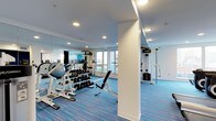 This single-floor fitness facility offers a thoughtfully designed workout environment that balances functionality with a clean, modern aesthetic. The space is anchored by vibrant blue striped flooring that adds energy and visual interest throughout the room. Recessed ceiling lighting provides consistent, even illumination across the entire area, complementing the natural light that streams in through large windows along one wall, offering views of the surrounding exterior.

A full-length mirror wall on one side visually expands the space and allows users to monitor their form during exercise. The facility is well-stocked with a comprehensive range of equipment, including a dumbbell rack with an organized selection of free weights, an adjustable weight bench, a cable pulley machine, and a multi-function strength training tower. A stability ball and cardio equipment complete the setup, catering to a variety of fitness routines. A wall-mounted flat-screen display adds a media element to the space. Structural white columns are integrated into the layout, adding an architectural detail while the crisp white walls and ceiling maintain a bright, open atmosphere throughout.