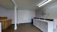 This single-floor laundry facility is a functional, communal space designed to accommodate residents' laundering needs. The room is finished with white-painted walls and features a concrete floor, giving it a clean, utilitarian appearance. Overhead fluorescent lighting provides ample illumination throughout the space.

Along one wall, a row of top-loading washing machines and dryers are neatly arranged side by side, accompanied by a wooden shelf above for placing laundry supplies. A wall-mounted air conditioning unit is positioned in the upper corner, ensuring comfortable temperatures during use. Instructional notices are posted on the walls, offering guidance for proper machine operation.

A structural column is present near the entrance, and the open floor layout provides sufficient room to move freely while loading and unloading machines. A waste bin is conveniently placed near the appliances. The space is straightforward and practical, prioritizing functionality with its streamlined design and essential amenities for an efficient laundry experience.