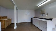 This single-floor laundry facility is a functional, communal space designed to accommodate residents' laundering needs. The room is finished with white-painted walls and features a concrete floor, giving it a clean, utilitarian appearance. Overhead fluorescent lighting provides ample illumination throughout the space.

Along one wall, a row of top-loading washing machines and dryers are neatly arranged side by side, accompanied by a wooden shelf above for placing laundry supplies. A wall-mounted air conditioning unit is positioned in the upper corner, ensuring comfortable temperatures during use. Instructional notices are posted on the walls, offering guidance for proper machine operation.

A structural column is present near the entrance, and the open floor layout provides sufficient room to move freely while loading and unloading machines. A waste bin is conveniently placed near the appliances. The space is straightforward and practical, prioritizing functionality with its streamlined design and essential amenities for an efficient laundry experience.