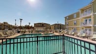 Located at 14510 Lemoyne Blvd in Biloxi, Mississippi, this residential community offers a welcoming Gulf Coast living experience defined by its bright, open surroundings and well-appointed shared amenities. The property's exterior showcases warm yellow stucco-finished multi-story buildings accented with white trim and private balconies, creating a cheerful and cohesive architectural aesthetic that is characteristic of coastal Southern design.

At the heart of the community is a generously sized outdoor swimming pool with clear turquoise water, serving as a central gathering point for residents. The pool deck is outfitted with numerous lounge chairs and seating areas, providing ample space for sunbathing and relaxation beneath the expansive blue sky. A white fence perimeter neatly defines the pool area, while black iron railings along elevated walkways offer safe and scenic vantage points overlooking the amenity space.

Mature palm trees and tasteful landscaping are visible throughout the grounds, enhancing the tropical, resort-like atmosphere of the community. Decorative street-style lamp posts add a charming touch to the outdoor environment. The single-floor unit layout ensures convenient, accessible living, while the overall design and shared outdoor spaces reflect a community built for comfort and enjoyment in the heart of the Mississippi Gulf Coast.