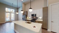 This contemporary single-floor residence is designed with a clean, modern aesthetic and thoughtful attention to detail throughout. The open-concept kitchen serves as the heart of the space, anchored by a generous white quartz island with an integrated sink and illuminated by a trio of stylish globe pendant lights. Cabinetry blends white upper shaker-style cabinets with warm wood-toned lower cabinets, creating a refined two-tone palette. A gray tile backsplash complements the full suite of stainless steel appliances, including a French door refrigerator, electric range, and built-in microwave. The soaring ceilings amplify the sense of openness, while light wood-tone flooring flows seamlessly throughout the living areas, adding warmth and continuity. A built-in wall niche provides functional display or storage space, and a closet near the entryway adds practical organization. The bathroom is finished with large-format tile flooring, a wood-toned vanity with white quartz countertop, a framed mirror with Hollywood-style lighting, and a glass-enclosed tiled shower. Natural light filters in through windows, further brightening the clean, neutral palette that defines the overall ambiance of this well-appointed space.