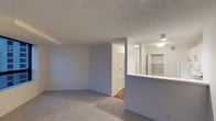 This single-floor space presents a clean, functional layout centered around an open living area finished with wall-to-wall carpeting in a neutral tone. The textured ceiling adds a classic touch overhead, while the soft grey walls contribute to a calm, understated atmosphere throughout.

A defining architectural feature is the partial wall with a pass-through counter, which visually separates the living space from an adjacent area while maintaining an open, connected feel. This design element also provides a practical surface for everyday use.

Built-in shelving is visible beyond the pass-through, offering organized storage and display space. The shelving unit is well-lit, enhancing both functionality and visual appeal in that section of the unit.

Further into the space, a doorway leads to what appears to be a bathroom, featuring tile flooring that contrasts with the carpeted main area. Additional doorways suggest further rooms or closet access, contributing to a practical and well-organized floor plan.

Flush-mount ceiling lights provide consistent illumination throughout, ensuring the space remains bright and welcoming. Overall, the interior offers a straightforward, move-in-ready environment with a neutral palette suited to a variety of furnishing styles.