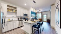 This single-floor residence showcases a well-designed open-concept layout that seamlessly connects the kitchen, dining, and living areas. The kitchen is a standout feature, equipped with stainless steel appliances including a French door refrigerator, electric range, microwave, and dishwasher. Light-toned cabinetry with open display shelving pairs with granite countertops, extending to a central island with bar seating and pendant lighting overhead. Wood-look flooring runs throughout the main living areas, adding warmth and continuity.

The living room is distinguished by a bold teal accent wall and large windows with access to an outdoor balcony, allowing generous natural light. Crown molding and a ceiling fan contribute to the room's polished character. Two bedrooms are present, each featuring ceiling fans, plush carpeting, and large windows that bring in natural views. The bathrooms are finished with granite-topped vanities, wood-look flooring, and ample storage. A bathroom conveniently positioned near the bedroom includes access to a closet. Throughout, the design balances modern finishes with vibrant color accents, creating a cohesive and inviting atmosphere.