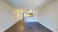 This single-floor unit offers an open and functional layout highlighted by dark hardwood-style flooring that extends throughout the main living area, creating a warm and cohesive aesthetic. The living space is generously proportioned, with clean white walls and a textured ceiling that add a classic, understated character to the interior.

A defining feature of the space is the pass-through kitchen window, which connects the kitchen to the main living area, promoting an open feel while maintaining a distinct cooking zone. The kitchen is equipped with warm wood-toned cabinetry and a ceiling-mounted flush light fixture that provides bright, focused illumination. A pendant light fixture is visible in the adjacent area, adding a subtle decorative touch.

The layout flows naturally from the main living zone into a secondary area, which benefits from a vertical window treatment that allows natural light to filter into the space. A flush-mount ceiling fixture provides additional overhead lighting. The neutral color palette and open floor plan make the space highly adaptable to a variety of furnishing styles and functional arrangements.