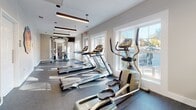 Located at Laurel Village in Henrico, Virginia, this well-appointed fitness center offers residents a comprehensive and modern exercise facility on a single level. The space is thoughtfully designed with a clean, contemporary aesthetic, featuring neutral warm-toned walls, durable speckled rubber flooring, and stylish pendant and linear suspended lighting that creates a bright, motivating atmosphere.

The cardio zone is well-equipped with multiple Matrix-brand treadmills, elliptical trainers, and upright exercise bikes, all arranged along a wall of large windows that flood the space with natural light and offer views of the surrounding community. The dedicated strength training area features a multi-functional cable machine system, along with additional floor space and rubber matting for free-form workouts and stretching.

A standout architectural element is the full-height glass partition wall with sleek black metal framing, which elegantly separates the fitness area from an adjacent lounge or leasing office space. This transparent division maintains an open, airy feel while defining distinct functional zones. The neighboring room, visible through the glass, features warm wood-look flooring, pendant lighting, and comfortable seating, adding to the overall welcoming character of the facility. Safety equipment, including a fire extinguisher and first aid provisions, is also conveniently accessible throughout the space.