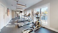 Located at Laurel Village in Henrico, Virginia, this well-appointed fitness center offers residents a comprehensive and modern exercise facility on a single level. The space is thoughtfully designed with a clean, contemporary aesthetic, featuring neutral warm-toned walls, durable speckled rubber flooring, and stylish pendant and linear suspended lighting that creates a bright, motivating atmosphere.

The cardio zone is well-equipped with multiple Matrix-brand treadmills, elliptical trainers, and upright exercise bikes, all arranged along a wall of large windows that flood the space with natural light and offer views of the surrounding community. The dedicated strength training area features a multi-functional cable machine system, along with additional floor space and rubber matting for free-form workouts and stretching.

A standout architectural element is the full-height glass partition wall with sleek black metal framing, which elegantly separates the fitness area from an adjacent lounge or leasing office space. This transparent division maintains an open, airy feel while defining distinct functional zones. The neighboring room, visible through the glass, features warm wood-look flooring, pendant lighting, and comfortable seating, adding to the overall welcoming character of the facility. Safety equipment, including a fire extinguisher and first aid provisions, is also conveniently accessible throughout the space.