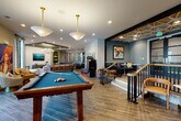 This well-appointed amenity lounge is thoughtfully designed to offer a diverse range of entertainment and social spaces within a single, open floor plan. The layout flows seamlessly between distinct zones, each with its own character while maintaining a cohesive contemporary aesthetic throughout.

Wood-look tile flooring laid in a herringbone pattern anchors the space, complementing the warm tones of the leather bar stools lining an extended counter-height bar. A decorative metal lattice partition elegantly defines zones without fully enclosing them, preserving an open, airy feel. Recessed lighting is supplemented by statement chandeliers and pendant fixtures, including a bold brass-and-glass pendant above the pool table area.

The gaming zone features a full-size billiards table as its centerpiece, accompanied by an illuminated shuffleboard table. A lounge area offers a generously sized sectional sofa beneath a tray ceiling accented with teal paint detail and a contemporary chandelier. Large windows and glass doors allow natural light to filter in while providing views of the surrounding exterior.

Bold, colorful artwork is displayed throughout, contributing to the energetic and modern atmosphere. Additional seating vignettes and a built-in shelving area complete this vibrant, multifunctional gathering space.