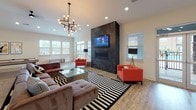 Located at 810 Eden Ave in Fort Mill, SC, this single-floor community space combines contemporary design with functional amenities. The expansive open-plan layout is anchored by a dramatic living lounge area showcasing a floor-to-ceiling dark tile fireplace surround with a wall-mounted flat-screen TV and a sleek linear gas fireplace. An eye-catching sculptural chandelier with exposed Edison bulbs adds an artistic focal point overhead, while warm wood-look flooring and bold black-and-white striped area rugs create a striking visual contrast throughout. Large windows fitted with plantation shutters allow generous natural light to fill the space. The lounge area comfortably accommodates ample seating, including a spacious sectional sofa and vibrant accent chairs, alongside a glass coffee table. A pool table and dining area are also integrated into the open layout, making this an ideal gathering destination. The kitchen features crisp white cabinetry, granite countertops with a generous island, a dark tile backsplash, stainless steel appliances including a French door refrigerator and dishwasher, and a dedicated sink station. Glass partition doors connect the main areas, maintaining an open, airy feel while delineating distinct zones within this well-appointed, versatile community space.
