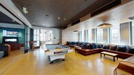 This single-floor amenity space is designed to accommodate a wide range of social, recreational, and wellness activities within a cohesive, contemporary aesthetic. Polished concrete floors flow throughout, unifying the various zones and lending an industrial yet refined character to the overall environment.

The central lounge area serves as the heart of the floor, featuring a striking billiard table as its focal point, surrounded by generous sectional seating in rich brown leather accented with deep teal cushions. Large windows along one wall admit abundant natural light and frame urban views. A bold, angular architectural beam cuts diagonally through the lobby transition area, adding a dramatic sculptural element, while a geometric wood-paneled accent wall further elevates the visual interest.

The communal kitchen is outfitted with a substantial marble-faced island, dark countertops, and a herringbone tile backsplash, complemented by floor-to-ceiling windows. An adjacent dining area features a long reclaimed wood table beneath a distinctive ring-style pendant chandelier.

The fitness facilities include a cardio room lined with treadmills and city-view windows, and a dedicated cycling studio with mirrored walls and a ballet barre. Flexible event space with high-top seating and draped curtain dividers rounds out the amenities.