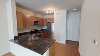 This single-floor space centers around a well-appointed kitchen that serves as the heart of the layout. The kitchen features warm, medium-toned wood cabinetry providing generous storage, complemented by dark granite countertops that extend to a peninsula with a built-in stainless steel sink — ideal for casual dining or meal prep. A full suite of stainless steel appliances, including a refrigerator, electric range, and over-the-range microwave, adds a modern, functional touch. A track lighting fixture overhead ensures the kitchen is well-lit throughout. The flooring throughout the space is a light hardwood-style material that adds warmth and continuity, connecting the kitchen to the entryway. The entry area is clearly defined, featuring a solid white door with multi-point hardware, offering a clean and secure transition into the unit. The walls are finished in a neutral white tone, keeping the space feeling bright and open. The overall design balances practicality with modern aesthetics, making efficient use of the available layout while maintaining a clean, cohesive appearance.