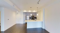 This single-floor residence offers a thoughtfully designed open-concept layout that seamlessly connects the kitchen and living areas. The kitchen is a standout feature, showcasing gray shaker-style cabinetry, a bold dark subway tile backsplash, and a full suite of stainless steel appliances, including a refrigerator, microwave, and range. A white peninsula with a built-in sink extends into the living space, providing additional prep space and creating a natural division between the kitchen and the open living area. Pendant lighting hangs above the peninsula, adding warmth and a modern touch. Recessed lighting and track lighting throughout the ceiling ensure the space is well-lit and versatile. The rich, dark hardwood flooring runs throughout the unit, grounding the bright white walls and cabinetry with a warm contrast. The entry door is conveniently positioned near the kitchen, with clean sightlines across the entire floor plan. The overall ambiance is clean, contemporary, and functional, making this a well-appointed space suited for modern living.