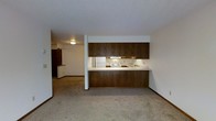 This single-floor space is designed with an open, functional layout that seamlessly connects the kitchen and living areas. The kitchen is a standout feature, showcasing rich, dark wood cabinetry that extends from floor to ceiling, providing ample storage. A white electric range, refrigerator, and a double sink are built into the kitchen, which also features a wraparound breakfast bar with a light-toned countertop — ideal for casual dining or additional prep space. The kitchen is partially open to the living area, separated by a low half-wall that maintains a sense of openness while offering a defined boundary between the two spaces. The living area is carpeted in a neutral beige tone, contributing to a warm and understated ambiance throughout. A textured ceiling adds a classic touch consistent with the overall design. A doorway visible in the background hints at additional rooms beyond, suggesting a thoughtful floor plan that extends further into the unit. Flush-mount ceiling lighting provides even illumination, and the neutral wall palette keeps the space feeling bright and adaptable. Overall, the layout is practical and approachable, suited for everyday living.