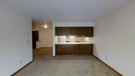 This single-floor space is designed with an open, functional layout that seamlessly connects the kitchen and living areas. The kitchen is a standout feature, showcasing rich, dark wood cabinetry that extends from floor to ceiling, providing ample storage. A white electric range, refrigerator, and a double sink are built into the kitchen, which also features a wraparound breakfast bar with a light-toned countertop — ideal for casual dining or additional prep space. The kitchen is partially open to the living area, separated by a low half-wall that maintains a sense of openness while offering a defined boundary between the two spaces. The living area is carpeted in a neutral beige tone, contributing to a warm and understated ambiance throughout. A textured ceiling adds a classic touch consistent with the overall design. A doorway visible in the background hints at additional rooms beyond, suggesting a thoughtful floor plan that extends further into the unit. Flush-mount ceiling lighting provides even illumination, and the neutral wall palette keeps the space feeling bright and adaptable. Overall, the layout is practical and approachable, suited for everyday living.