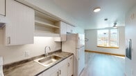 Located at 12370 SE Ash St in Portland, Oregon, this single-floor unit offers a clean, efficiently designed layout with a bright, modern aesthetic throughout. The kitchen is well-appointed with white cabinetry, open shelving, a double-basin stainless steel sink, dark laminate countertops, a coil-burner electric range, and a full-size refrigerator — all finished in a cohesive white palette that keeps the space feeling open and functional. Light wood-tone flooring runs throughout the main living areas, complemented by warm wood baseboards that add a touch of natural contrast to the neutral white walls. The open living area benefits from natural light streaming through a large window with views of the surrounding neighborhood's greenery and architecture, and is equipped with a ceiling fan for year-round comfort. A built-in wall shelf provides additional storage or display space in the living area. The bathroom features a full tub-shower combination, a vanity with a dark countertop and undermount sink, a large mirror, and clean white walls, offering a practical and tidy space. The floorplan, as seen in the overhead layout view, also includes an accessible bathroom area with grab bars and a roll-in shower, making this unit suitable for a range of accessibility needs.