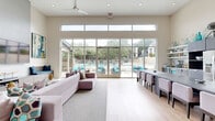 This generously sized single-floor facility showcases a contemporary design centered around an open, light-filled lounge area. Soaring ceilings and expansive floor-to-ceiling glass walls with clerestory windows above flood the interior with natural light while offering direct views of an outdoor pool and landscaped surroundings. Light hardwood flooring runs throughout, reinforcing the clean, modern aesthetic. The lounge features large, comfortable sectional seating positioned around a wall-mounted television, with frosted glass partition walls creating a refined separation between spaces without sacrificing openness.

The hallway connects the main areas with a seamless flow, incorporating glass-paneled doors that maintain visual continuity. The dining room provides a functional gathering space with multiple table configurations, ceiling fans, elevated clerestory windows, and dual wall-mounted displays suited for presentations or shared viewing. The two offices offer versatile work environments; one features vibrant accent walls, built-in shelving, workstations, and a casual round table, while the other is a clean, minimalist room with light walls and hardwood floors. A covered outdoor dining area with a ceiling fan, glass-top table, and wicker seating completes the facility's amenities.