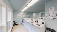 This shared laundry facility is a single-floor space designed for efficiency and ease of use. The room is finished with light blue walls and neutral vinyl flooring, creating a clean and utilitarian atmosphere. Overhead fluorescent lighting illuminates the entire space evenly, ensuring good visibility throughout.

The facility is equipped with two distinct appliance zones. Along one side, a row of top-loading washing machines is arranged in a line, accompanied by front-loading washers at one end. The opposite wall features stacked dryer units, maximizing the use of vertical space. All machines appear to be modern commercial-grade units with digital controls. Informational signage, including laundry tips and usage instructions, is posted on the walls for user guidance.

Seating is available via folding chairs placed at either end of the room, offering a place to wait during laundry cycles. A set of double doors with an illuminated exit sign provides the main entry and exit point, while a window adjacent to the doors allows a view of greenery outside, introducing a small amount of natural light. The overall layout is straightforward and practical, prioritizing functionality and accessibility for shared use.