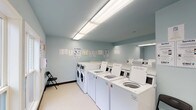 This shared laundry facility is a single-floor space designed for efficiency and ease of use. The room is finished with light blue walls and neutral vinyl flooring, creating a clean and utilitarian atmosphere. Overhead fluorescent lighting illuminates the entire space evenly, ensuring good visibility throughout.

The facility is equipped with two distinct appliance zones. Along one side, a row of top-loading washing machines is arranged in a line, accompanied by front-loading washers at one end. The opposite wall features stacked dryer units, maximizing the use of vertical space. All machines appear to be modern commercial-grade units with digital controls. Informational signage, including laundry tips and usage instructions, is posted on the walls for user guidance.

Seating is available via folding chairs placed at either end of the room, offering a place to wait during laundry cycles. A set of double doors with an illuminated exit sign provides the main entry and exit point, while a window adjacent to the doors allows a view of greenery outside, introducing a small amount of natural light. The overall layout is straightforward and practical, prioritizing functionality and accessibility for shared use.