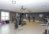 This single-floor fitness center is designed to provide a comprehensive workout environment for users of all fitness levels. The space is finished with deep charcoal walls that lend a focused, energetic ambiance, while a light-colored floor provides a clean contrast and ample visibility throughout the room.

At the heart of the gym sits a multi-functional cable and weight machine, offering a wide range of strength training exercises in one central station. Flanking this unit are adjustable benches, allowing for versatile use of the equipment. Along the right wall, a well-organized free weight rack holds a full set of kettlebells and dumbbells in graduated sizes, accompanied by a wall-mounted medicine ball rack stocked with a colorful array of weighted balls to support dynamic training routines.

Natural light filters in through a window on the far wall, brightening the space during daytime hours. Two ceiling fans are strategically positioned overhead to maintain air circulation and comfort during intense workouts. A large mirror along one wall enhances the sense of space while allowing users to monitor their form. Motivational fitness imagery is displayed on the walls, reinforcing the gym's athletic atmosphere. An exit door provides convenient access, rounding out this practical and well-appointed fitness facility.