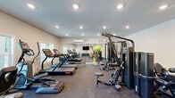 Located at 1515 Hogansville Rd in LaGrange, Georgia, this fitness center offers a comprehensive workout facility designed to support a wide range of exercise routines. The single-room space is thoughtfully laid out to maximize functionality while maintaining an open, accessible feel throughout.

The gym features a robust selection of cardio equipment, including multiple treadmills and an elliptical trainer, positioned along windowed walls that allow natural light to filter in and provide views of the surrounding greenery. A wall-mounted flat-screen television is available for entertainment during workouts.

Strength training is well-supported with a multi-station cable machine system, a variety of resistance machines, and a full rack of dumbbells spanning a wide weight range. Stability balls and additional accessories round out the free-weight area. A dedicated bench provides seating for targeted exercises.

The flooring consists of durable rubber gym matting, offering safety and comfort underfoot. A wall-mounted split air conditioning unit ensures the space remains climate-controlled year-round. Full-length mirrors along one wall enhance the sense of space and allow for proper form checks during exercise. A built-in dual water fountain station is conveniently located near the entrance, providing easy hydration access. Recessed lighting throughout keeps the space bright and well-illuminated.