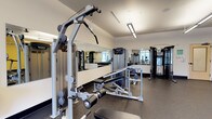This dedicated fitness center is thoughtfully designed to support a comprehensive workout experience. The space is outfitted with professional-grade equipment from leading brands, including Precor strength-training machines and True cardio equipment. Strength training options include a cable lat pulldown machine, a multi-functional weight bench with leg extension attachment, a functional trainer, and a dumbbell rack stocked with a full range of free weights. Cardio equipment, including rowing-style machines, is positioned along one side of the room. The floor is covered in durable, shock-absorbing rubber flooring in a speckled gray finish — a practical and industry-standard surface for high-traffic gym environments. Full-length mirrors line one wall, allowing users to monitor their form and making the space feel more open and expansive. Overhead, a series of linear LED fixtures and industrial-style pendant lights provide bright, even illumination throughout. Natural light filters in through windows visible in the mirror reflections. Ceiling fans contribute to air circulation, enhancing comfort during workouts. The overall aesthetic is clean, modern, and functional, creating an inviting environment suited for a variety of fitness routines.