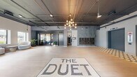 The Duet's ground-floor lobby in White Plains, NY, presents a thoughtfully designed residential common area that blends industrial aesthetics with contemporary elegance. The entrance is immediately distinguished by a signature mosaic tile floor inlay bearing "THE DUET" branding, light hardwood-style flooring, and dramatic exposed ceiling infrastructure with dark-painted ductwork and piping that lend an urban, loft-inspired character throughout.

The main lobby area features a striking multi-globe chandelier as a central focal point, complemented by patterned geometric accent walls in deep slate tones. A dedicated mailroom section houses a full bank of resident mailboxes alongside a smart parcel management system, conveniently positioned near the main entrance.

Flowing seamlessly from the lobby, the lounge and social area offers a curated mix of seating, including a rich leather sofa, café-style bistro chairs, and accent tables set atop a patterned area rug. Decorative arched glass display shelving and a kitchenette with dark cabinetry and a sink serve the social space, which is also furnished with booth-style dining seating beneath warm pendant lighting.

Large street-facing windows flood the space with natural light and offer views of the surrounding White Plains streetscape. Lush potted plants are thoughtfully placed throughout, softening the industrial design elements and adding a vibrant, welcoming ambiance to this well-appointed residential lobby.
