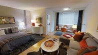 This single-floor unit showcases an efficient open-plan design that seamlessly integrates the living, dining, and sleeping areas into one cohesive space. The warm parquet wood flooring runs throughout, adding texture and visual continuity across the entire unit.

The sleeping area is positioned toward one end of the space, anchored by a full-sized bed dressed in dark bedding and complemented by a crisp white dresser topped with decorative accents and a framed mirror. A bedside table with a classic white lamp adds a soft ambient glow to the corner.

The living area centers around a round marble-topped coffee table, accented with a decorative bowl, and is flanked by a cushioned sofa adorned with warm orange throw pillows. A cowhide-patterned area rug grounds the seating arrangement, adding a stylish contrast to the wood floors.

Adjacent to the living space, a compact dining area features a round white table with modern orange molded chairs, bathed in natural light from a large window fitted with slatted blinds and dark curtain panels. A glimpse of a kitchenette is visible in the background, completing the functional layout of this well-appointed unit.