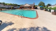 Located at 300 Pelham Rd in Greenville, South Carolina, this outdoor amenity space centers around a generously sized swimming pool with striking turquoise water, framed by a classic brick-edged coping and a wide surrounding concrete deck. The pool area is well-appointed for outdoor leisure, featuring multiple lounge chairs positioned along the perimeter for sunbathing, as well as shaded patio seating areas with blue market umbrellas and wicker-style tables and chairs — ideal for relaxing or socializing. A charming single-story clubhouse anchors the background of the space, constructed with warm brick and lap siding, a pitched roof, and a chimney, lending a welcoming residential character. Large windows along the clubhouse facade allow natural light to flow in while offering views of the pool. The entire area is enclosed by a decorative black wrought-iron fence, adding both security and visual appeal. Lush, mature landscaping — including evergreen shrubs, flowering plants in large terracotta pots, and tall shade trees — lines the fence line and softens the hardscape surroundings. The space enjoys abundant natural light under open skies, creating a bright and inviting outdoor retreat well-suited for warm-weather enjoyment.