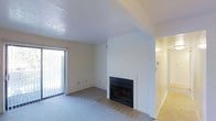 This single-floor residence presents a straightforward and functional layout designed for comfortable everyday living. The main living area is finished with wall-to-wall carpeting in a neutral tone, creating a soft and cohesive foundation for the space. A built-in fireplace serves as a natural focal point, set against a crisp white wall with a tiled hearth surround that provides a clean, classic look. The fireplace adds both visual interest and a practical warming element to the room.

Natural light enters the living area through a sliding glass door fitted with vertical blinds, offering convenient access to an outdoor area while keeping the interior bright and airy. The white walls and light-toned ceiling, which features a subtle textured finish, contribute to the open and uncluttered feel of the space.

A hallway visible from the living area connects to additional rooms, suggesting a logical and easy-to-navigate floor plan. The transition areas appear to feature hard flooring, contrasting with the carpeted main living space. Overall, the unit presents a clean, move-in ready environment with simple, durable finishes suitable for a variety of living arrangements.