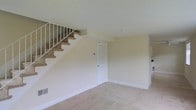 This two-story residential space presents a straightforward and functional layout designed for comfortable living. The main level features a carpeted open living area finished with neutral beige walls and white trim, creating a clean and understated aesthetic throughout. A carpeted staircase with a white-railed banister connects the two floors, positioned along one wall to preserve the flow of the main living space. Beneath the staircase, a white interior door provides access to additional storage or utility space, maximizing the practical use of the floor plan. The flooring is finished with consistent wall-to-wall carpet in a light neutral tone, contributing to a cohesive and simple interior palette. Recessed ceiling fixtures provide overhead lighting, while baseboards and trim throughout are kept in white, complementing the overall neutral color scheme. The space feels open and uncluttered, offering a blank canvas that can be adapted to a variety of furnishing styles. The second floor, accessible via the staircase, extends the living footprint and adds versatility to the overall layout of this two-level home.