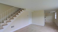 This two-story residential space presents a straightforward and functional layout designed for comfortable living. The main level features a carpeted open living area finished with neutral beige walls and white trim, creating a clean and understated aesthetic throughout. A carpeted staircase with a white-railed banister connects the two floors, positioned along one wall to preserve the flow of the main living space. Beneath the staircase, a white interior door provides access to additional storage or utility space, maximizing the practical use of the floor plan. The flooring is finished with consistent wall-to-wall carpet in a light neutral tone, contributing to a cohesive and simple interior palette. Recessed ceiling fixtures provide overhead lighting, while baseboards and trim throughout are kept in white, complementing the overall neutral color scheme. The space feels open and uncluttered, offering a blank canvas that can be adapted to a variety of furnishing styles. The second floor, accessible via the staircase, extends the living footprint and adds versatility to the overall layout of this two-level home.