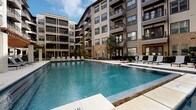This outdoor pool area is designed as a full-service amenity space, combining architectural elegance with functional leisure. The centerpiece is a generously sized swimming pool with clear blue water, finished with stone-accented edges and surrounded by a wide pool deck furnished with lounge chairs. The pool is framed on multiple sides by a multi-story building featuring balconies, large windows, brick and neutral-toned exterior cladding, and modern architectural detailing that creates an enclosed, resort-like atmosphere.

Adjacent to the pool is a covered terrace supported by substantial natural stone columns, providing a shaded retreat from direct sunlight. The terrace features large-format tile flooring in warm, earthy tones and is equipped with ceiling fans to promote airflow. Glass panel railings along the terrace perimeter maintain open sightlines to the pool and surrounding greenery.

The pool entrance area includes a gated access point with decorative metal fencing, lush landscaping, and string lights overhead, adding a welcoming ambiance. Seating throughout the space includes stylish black metal and woven outdoor chairs suited for both relaxed lounging and casual socializing. The overall design balances modern materials with natural stone accents, creating a polished, upscale outdoor environment.
